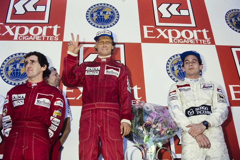 Niki Lauda, newly-crowned World Champion by finishing in 2nd position, stands atop of the podium with three fingers showing for each title alongside Alain Prost, 1st position, and Ayrton Senna, 3rd position.